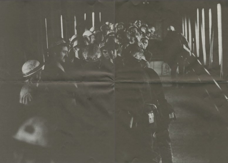 Black and white photograph depicting a group of industrial workers, predominantly wearing safety helmets, gathered in a corridor with limited lighting. Most workers appear engaged in conversation or standing attentively, while a few individuals are positioned towards the front with their backs to the camera. The setting suggests an industrial environment, emphasizing the historical context and working conditions typical of mid-20th century industry.