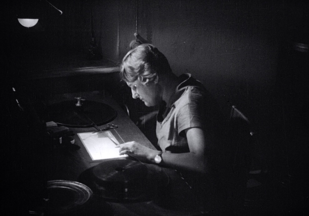 A person seated at a dimly lit desk is focused on examining or editing film strips. The scene includes film reels, a splicing machine, and a small desk lamp illuminating the workspace. The monochromatic image emphasizes the intricate details of the editing process.