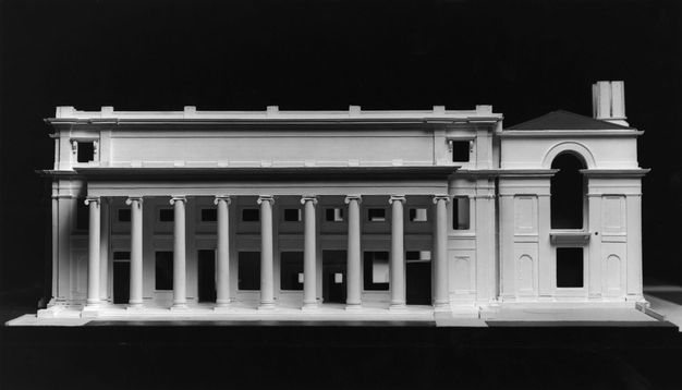 Black and white image of an architectural model featuring a neoclassical building with a row of 9 evenly spaced columns at the front. The background is dark.
