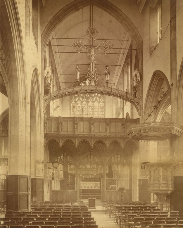 Sepia-toned photograph of a Gothic-style church interior. The image showcases the nave, featuring a high-vaulted ceiling and intricately carved wooden rood screen adorned with statues. A large, ornate cross is suspended above. Rows of wooden pews are visible, facing towards the altar, behind which is a stained glass window. A pulpit is to the right.