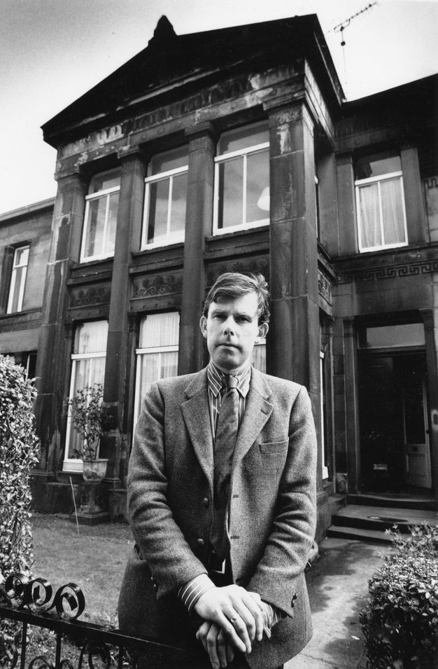 Black-and-white portrait of Gavin Stamp standing in front of a historic stone-fronted house with large sash windows and Corinthian-style pilasters, dressed in a tweed jacket and tie.