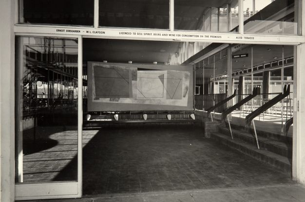Black and white photograph of an architectural interior featuring a modernist artwork displayed prominently on the wall. The room is characterized by large glass windows, which allow natural light to illuminate the space, creating reflections and shadows on the tiled floor. Geometrically designed ramps lead up from the tiled area, and signage indicates a licensed premise for the sale of spirits, beer, and tobacco. The artistic piece consists of abstract geometric shapes.