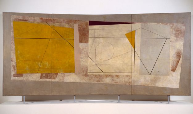 Abstract painting featuring geometric shapes and lines on a multi-paneled surface. The composition includes a large yellow area on the left with intersecting black lines forming various polygons. The right side shows a beige background with a complex arrangement of linear elements and a partial circle. Muted earthy tones, including browns and grays, form the backdrop, lending a textured and layered appearance. The artwork is displayed on a white wall and mounted on a stand.