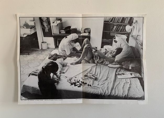 Black and white photograph depicting a domestic scene in a bedroom. Central focus is on a bed where a woman lies propped up against pillows, holding an infant. A person in medical attire attends to the woman, suggesting a recent birth. On the bed, a child is seen playing with Lego bricks. The room is furnished with shelves containing books, a lamp, scattered items on a desk, and boxes, creating a lived-in atmosphere. The image captures a candid moment of postpartum care and family interactions within
