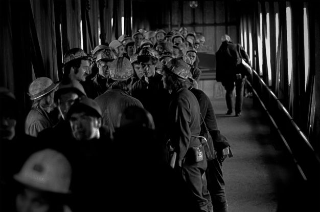 A group of miners, wearing helmets and work attire, gather in a narrow, enclosed walkway made of metal and wood at Kellingley Colliery, Yorkshire. They appear to be engaged in discussion, possibly regarding union matters. The confined space and expressions suggest a serious atmosphere. Light streams in from the right side through the structure‚Äôs windows, creating contrasting shadows. This photograph, captured by Robert Golden, provides insight into the social dynamics and working conditions within the mining industry.