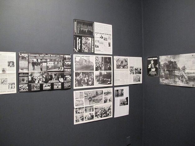 The image depicts a corner of a gallery or exhibition space showcasing a collection of historical newspaper clippings and photographs arranged on a dark gray wall. The display includes various articles and images, including a prominent headline reading "Community under attack: Brick Lane 1978," and another composition titled "Catholic West Belfast." The materials appear to document social and political events, reflecting community struggles and cultural narratives. This visual presentation is likely part of an exhibit focusing on historical socio-political contexts.