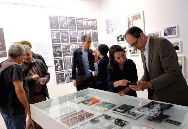 Visitors engage with exhibits at an art gallery or museum. Several individuals examine displayed photographs and documents in a glass case, while others discuss near a wall featuring various posters and framed images. The setting appears to be a presentation or special exhibition, focusing on the intersection of photography and its intended audience.