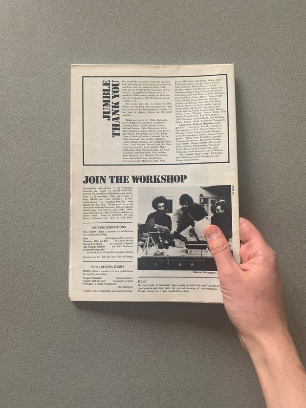 A hand holding an open black-and-white magazine page against a gray surface. The page features a column titled "Jumble Thank You" with a list of names, and a section titled "Join the Workshop" with information about exhibitions and workshops. A photograph is included showing three people engaged in a photography-related activity. The layout includes text blocks, headlines, and a photograph, typical of a magazine format. The image captures the vintage aesthetic and design of the publication.