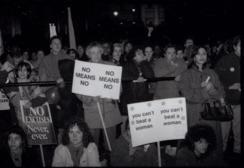Black and white photograph. A group of women of various ages hold three placards with the text "NO Excuses. Never, ever", "NO MEANS NO" and "you can't beat a woman". Four women sit on the ground, one has her face behind the "you can't beat a woman" sign. Many more people gather behind them at a railing, standing in front of a building.
