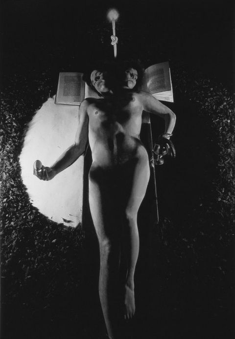 Black and white photograph. A naked person reclines - or is suspended by a small rope - across the centre of the image. They have two heads, each of which rests on an open book. Their body appears split into two, with a masculine, and a feminine, half. The hand on the left holds an egg over a semi-circle of light, while the right holds a wine glass over a semi-circle of darkness.