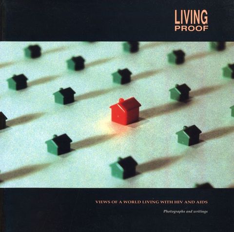 Cover of book. Colour photograph of several green plastic houses and one larger red house off-centre. Orange capitalised text in the upper-right corner "LIVING PROOF" and underneath the houses "VIEWS OF A WORLD LIVING WITH HIV AND AIDS" above the subtitle “Photographs and writings” in white text.