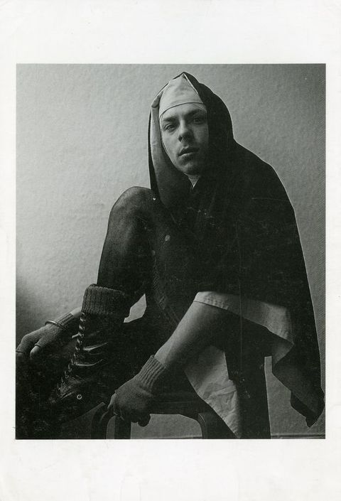 Black and white photograph. A person wearing a nun’s veil and wool, fingerless gloves is seated on a stool, looking out towards the camera against a plain white backdrop. One leg is bent, with a foot perched on the edge of the stool, revealing their thin, laddered tights, thick socks, and black, leather lace-up boots.