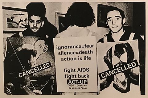 Black and white photograph. Three people stand side by side within a gallery space that has artworks hung on the wall. The person in the centre has their back towards us and wears a placard reading “ignorance = fear, silence = death, action is life, fight AIDS”. The two flanking him face towards us, both wearing placards emblazoned with the word “CANCELLED”.