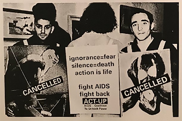 Black and white photograph. Three people stand side by side within a gallery space that has artworks hung on the wall. The person in the centre has their back towards us and wears a placard reading “ignorance = fear, silence = death, action is life, fight AIDS”. The two flanking him face towards us, both wearing placards emblazoned with the word “CANCELLED”.