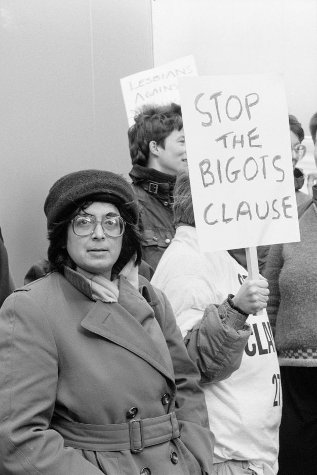 Black and white photograph. Person wearing a trench coat and wool hat stands on the left with their arm raised, holding up a protest sign displaying the uppercase text "STOP THE BIGOTS CLAUSE" in front of a group of protestors.   