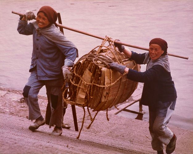 Colour photograph. Two Chinese women in working clothes and headscarves stand carrying a bamboo pole which holds folded brown cardboard boxes tied with rope. They smile while walking up the steps, appearing in front of a body of water in the background.