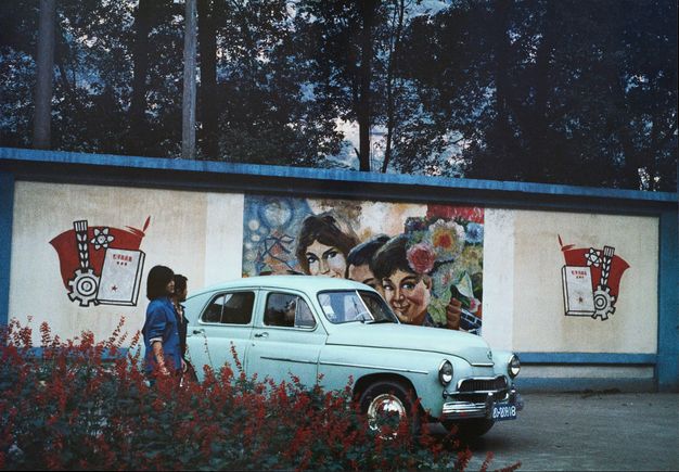 Colour photograph. A central light blue car is parked in front of a painted wall mural depicting three female smiling faces and flowers. Bordering this mural are two sets of red, white and black illustrations with Chinese symbols. Two people stand to the left of the vehicle; one gazes at the mural with trees visible behind the wall.