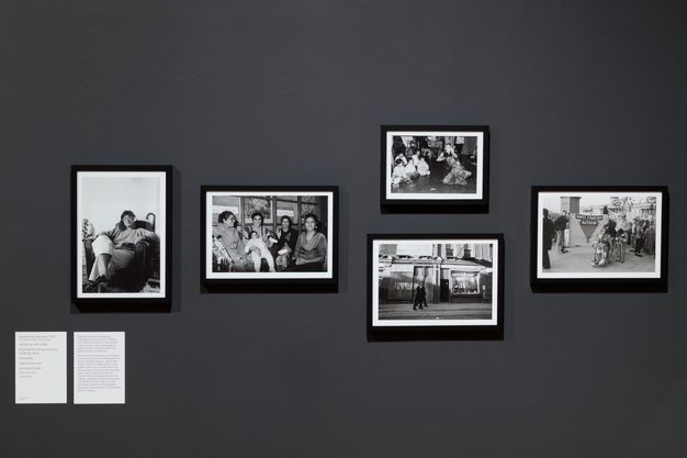 Installation photograph. Black and white. Five photographs in dark frames with light paper mounts are displayed on a grey wall. From left-to-right, the photographs depict an Indian person seated holding a pipe, an Indian family, an Indian dancer, two policemen walking past a store, and protestors outside a building holding an "Anti Facist Action" banner next to standing man and woman in a wheelchair.