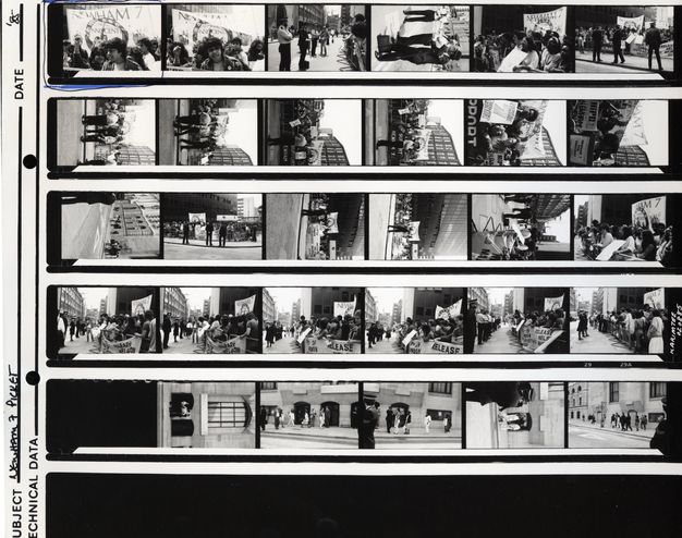 Black and white photograph. Series of 29 images on a contact sheet with protestors and police in a London street. Many feature a large banner with the text "NEWHAM 7" at the top and "Innocent" underneath. A banner is lifted behind a group of South Asian protestors raising their arms in the top-left image, which has been circled with blue ink.