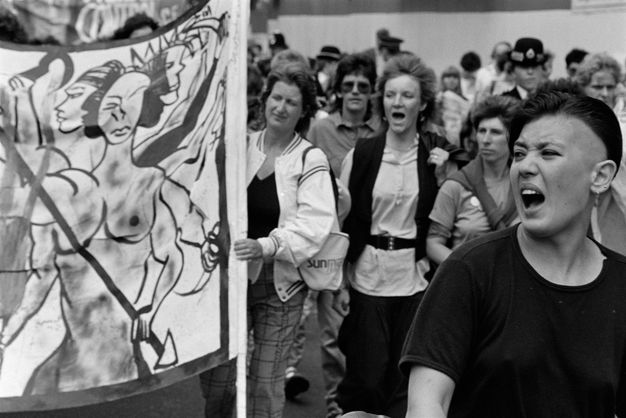 Black and white photograph. An open-mouthed woman stands looking past the left of the frame. A row in the middle ground includes three women protestors, the left woman holding a banner of three muscular naked figures, scythes and a spear. A uniformed policewoman stands in the background, looking towards the foreground.