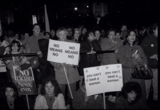Black and white photograph. A group of women of various ages hold three placards with the text "NO Excuses. Never, ever", "NO MEANS NO" and "you can't beat a woman". Four women sit on the ground, one has her face behind the "you can't beat a woman" sign. Many more people gather behind them at a railing, standing in front of a building.