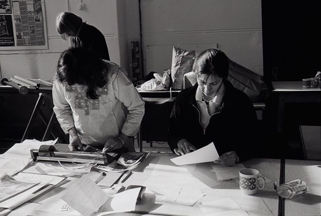 Black and white photograph. Three women work in an office or studio space. Two of them are looking down at a desk. One wears an Asian tunic, trimming photographic prints using a guillotine, while another on the right reads handwritten letters. Another woman stands working behind them.