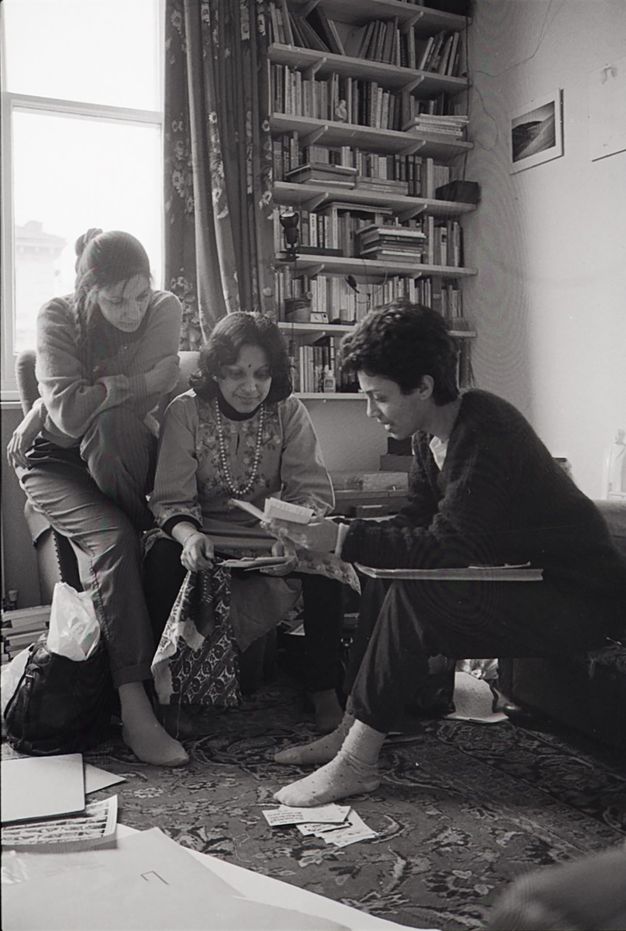 Black and white photograph. Three South Asian women look at postcards in the centre of a room. The middle, older woman sits on an armchair while a younger woman with plaited hair sits on the chair’s arm, her right leg touching an oriental-style carpet. A short-haired woman leans towards the others, her left foot covering postcards on the floor.