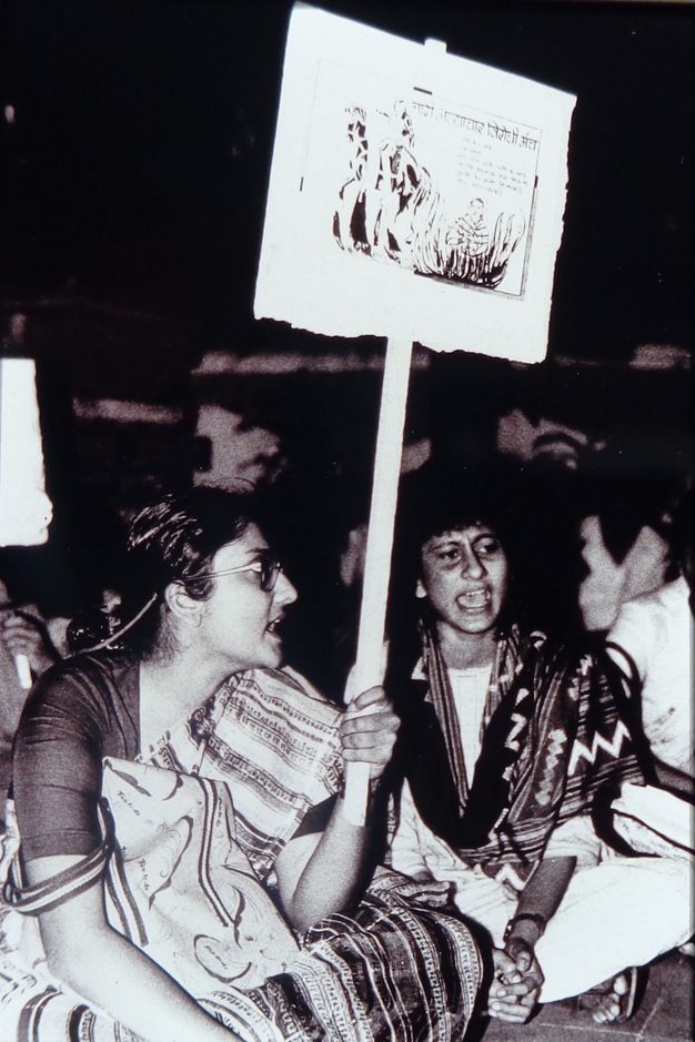 Black and white photograph. Two women wearing patterned saris across their shoulders sitting on the ground in protest. The woman on the left holds a sign on a wooden post that features an illustration of a burning widow. The background is dark and blurry.