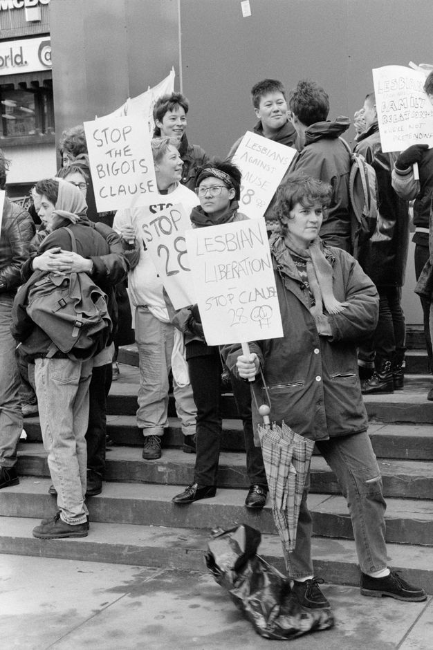 Black and white photograph. Group of 12 people standing on worn stone steps, holding six protest signs including the capitalised text "STOP THE BIGOTS CLAUSE", and "LESBIAN LIBERATION: STOP CLAUSE 28" featuring two female symbols. 