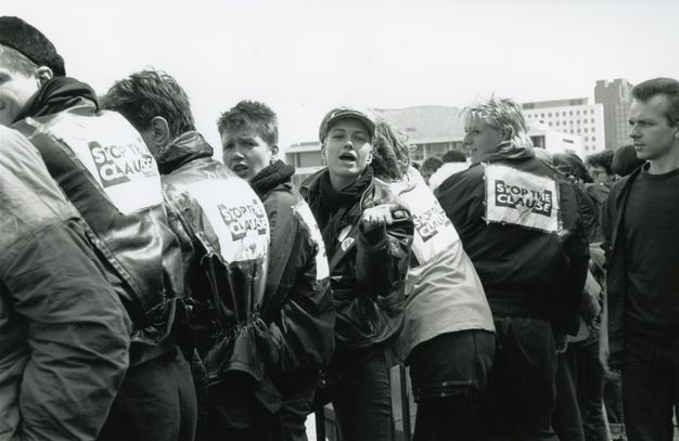 Black and white photograph. Group of six people wearing dark wax jackets featuring the "STOP THE CLAUSE" capitalised text and protest symbol; an inverted triangle intersecting the text. A seventh person looks leftwards towards the group, the majority of whom have their backs turned.