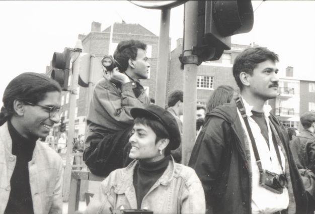 Black and white photograph. Four people standing in a rhombus arrangement at a traffic light. A central woman wearing a cloth hat and denim jacket talks to a man wearing glasses on the left. Another man stands to the right of her, a camera around his neck. The background street has apartments and shops.