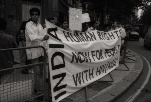 Black and white photograph. A large white cloth banner held by several protestors at the side of a busy road. The capitalised horizontal text "HUMAN RIGHTS NOW FOR PEOPLE WITH HIV/AIDS", the word "INDIA" appears vertically to the left of these words.
