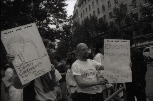 Black and white photograph. A bald man in a white t-shirt wearing glasses stands in the centre, holding a sign up. The sign's uppercase text "BLACK AIDS ACTION" and "PERSECUTION AND DISCRIMINATION WON'T STOP AIDS. EDUCATION WILL"