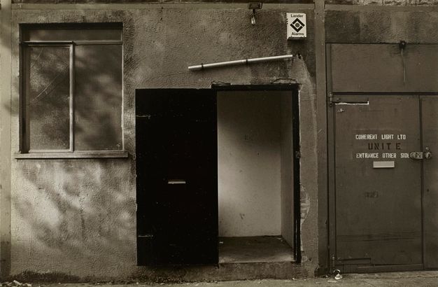 Black and white photograph. Close-up of a rendered concrete building and black door with a letterbox is open in the centre, near a window and security alarm box. Capitalised stencilled text reads "ENTRANCE OTHER SIDE".