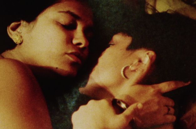 Colour film still. Close-up view of two medium-brown skin women's faces and shoulders reclined on a textured green wool blanket. The left woman closes her eyes, her right arm holding the other woman's neck, with one finger touching her counterpart's short hair. The second woman holds the other's gold bangled hand.