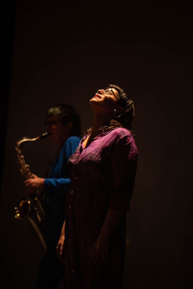 Performance art. Two people stand including a glasses-wearing woman wearing a buttoned floral silk pink dress in the centre and a saxophonist in a mid-blue suit playing behind her in this colour photograph.
