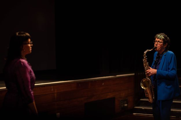 Performance art. Two women stand facing each other, the left woman wearing glasses and a fitted pink silk dress. A saxophone-playing woman with short hair and a mid-blue suit looks towards the first woman in front of a stage and steps in this colour photograph.