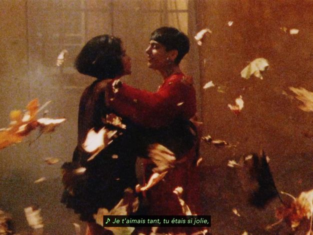 Colour film still. A woman wearing a dark red mesh dress with voluminous skirt, standing slightly left of centre with her right arm on another person who wears floaty crimson outfit. They dance surrounded by falling autumnal leaves against a window. French italicised subtitles "Je t'aimais tant, tu étais si jolie".