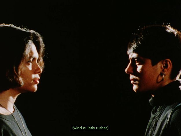 Colour film still. Two women stand at either side, looking into each other's brown eyes. One has a bob hairstyle, grey top and metallic gold necklace. The other wears shiny gold hoop earrings, short hair with a fringe and embellished grey polo neck jumper. Green subtitle text "wind quietly rushes" against a black background.