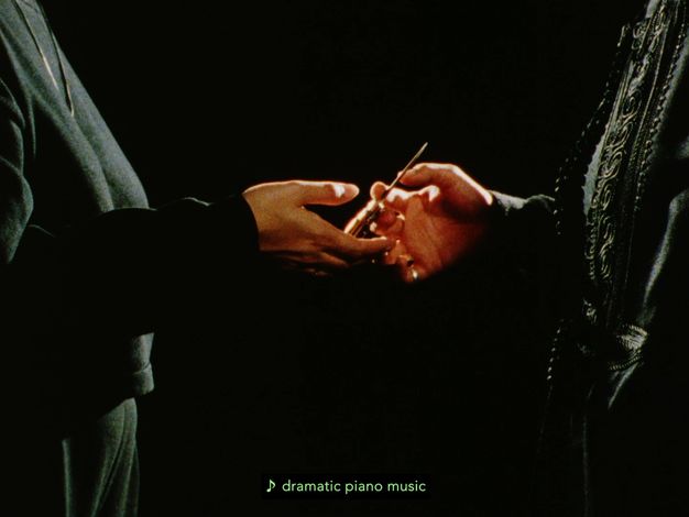 Colour film still. Two women shown from their shoulders down to their thighs, wearing grey clothing. The woman on the right has a shiny gold band ring on her right hand, passing a metallic object into the other's woman's hand. Green subtitles "dramatic piano music" against a black backdrop.