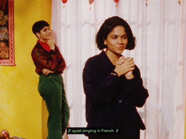 Colour film still. A woman standing in the foreground with her hands clasped, her golden jewellery shining. Behind her, a short-haired woman wearing a loose tapestry-style jumper smiles towards her. Green subtitles “quiet singing in French” appear over the room decorated with yellow walls, tapestry decorations and wooden instruments.