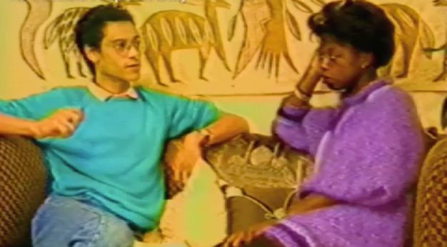 Video still. Two people in 1980s period attire sit on a sofa in conversation.