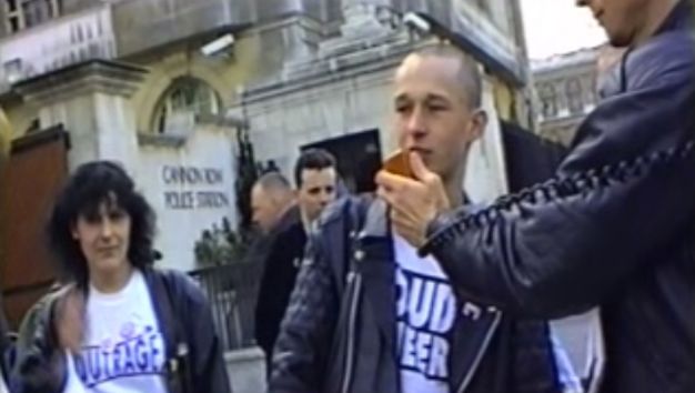 Film still. Two individuals leaving a police station wearing t-shirts with campaign slogans. Another individual extends a microphone to one of the people.