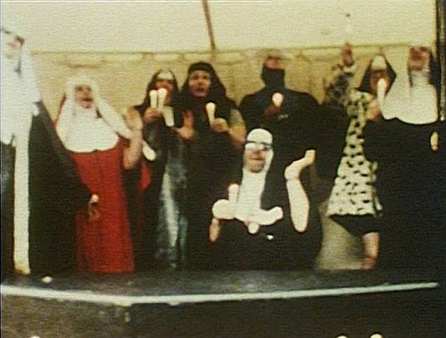 Colour film still. Seven people standing in a canvas tent singing and holding lit candles. One wears a red nun’s habit; the others are predominantly dressed in black and white habits. In front, a man wearing a black nun's habit waves his hand backwards, kneeling behind a black wooden coffin.
