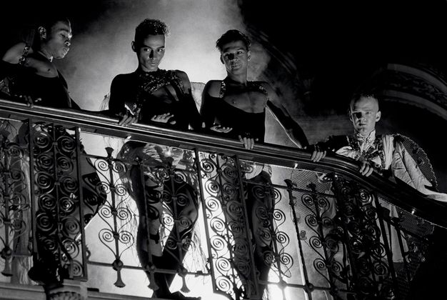 Black and white film still. Three men wearing matching black studded leather harnesses and draped shorts stand on a wrought iron balcony with curled metalwork, a fourth man on the right wears feathered wings and a cloth tunic. Clouds of smoke and ornate arches fill the background.