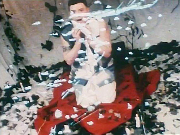 Colour film still. A man kneels on a red cloth wearing a black and white wool jacket and light trousers. White confetti falls from above inside a canavs tent, with trailing ivy in the background.
