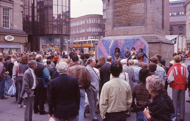 A black and white photograph of a performance of *The Last Blind Date Show* at Grey's Monument, Newcastle upon Tyne, 22 May 1992. The image shows a group of people gathered around a small stage, with a performer in the centre of the frame. The performer is wearing a white suit and is holding a microphone. The audience is visible in the background, with some people sitting on the ground and others standing. The monument is visible in the background, with a large crowd of people gathered around it. The sky is overcast, and the ground is wet, suggesting that it has recently rained.