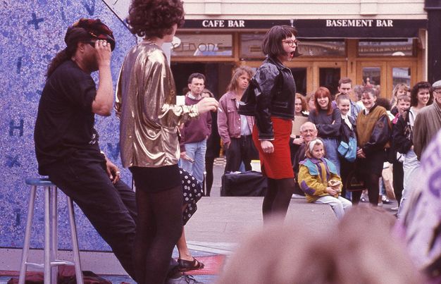 Performance art. A woman wearing a loose metallic blouse standing by a guy dressed in black clothes sitting on a stool. Another woman in the middle ground has a bob, leather jacket, fitted red mini skirt and glasses, her mouth opened. The on-street crowd laughs in the background of this colour photograph.