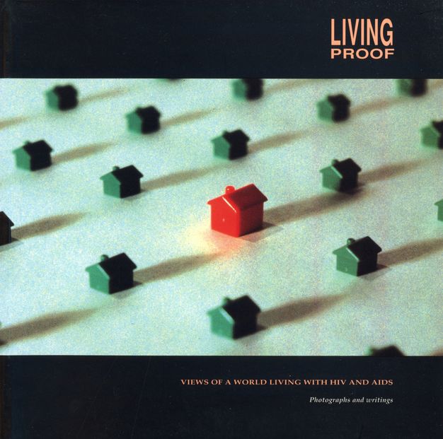 Cover of book. Colour photograph of several green plastic houses and one larger red house off-centre. Orange capitalised text in the upper-right corner "LIVING PROOF" and underneath the houses "VIEWS OF A WORLD LIVING WITH HIV AND AIDS" above the subtitle “Photographs and writings” in white text.