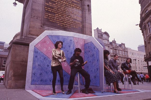 Performance art. A woman wearing a loose metallic gold blouse, and man in black baggy clothing are divided from two women and a man, all seated on stools by a screen. The two sides of the blue and pink screen have the letters "H", "I", "V" and the "+" symbol, and "A", "I", "D", "S" jumbled, against a town centre backdrop in this colour photograph.